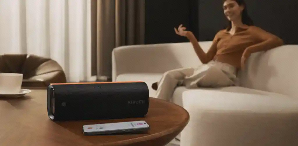 اسپیکر Xiaomi Sound Party و کنترل روی میز اسپیکر Xiaomi Sound Party روی میز چوبی کنار کنترل با فرد نشسته روی مبل روشن