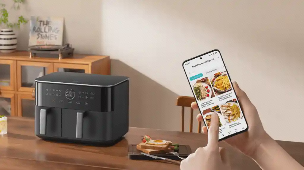 هواپز Xiaomi MAF-D1001 و برنامه دستور غذا هواپز Xiaomi MAF-D1001 روی میز کنار گوشی در دست فرد با تصاویر دستور غذا روی نمایشگر