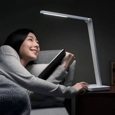 چراغ Mijia Table Lamp Lite کنار مبل برای مطالعه شبانه زن در حال مطالعه کتاب روی مبل کنار چراغ روشن Mijia Table Lamp Lite و دست روی پایه