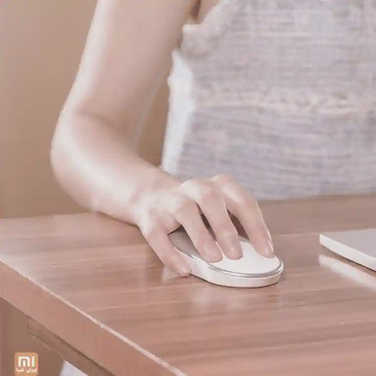 دست روی موس تخت و روشن Mi Portable Mouse روی میز چوبی کنار دفتر